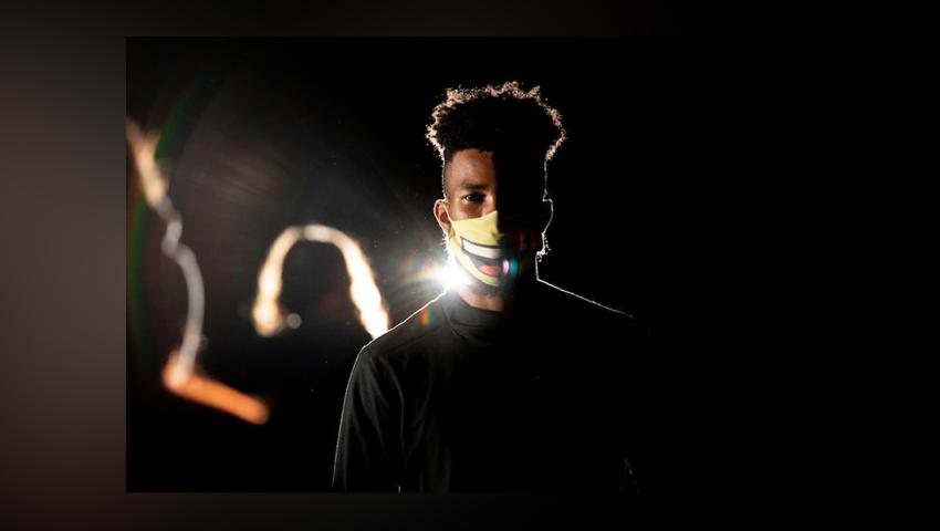 Student wearing white mask and dark shirt faces camera, backlight with other students onstage blurred in background
