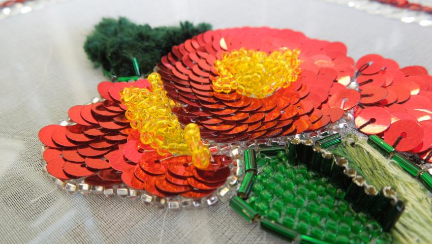 A thin white netting fabric is stretched over a frame and adorned with bright red sequins, yellow glass beads, and metallic green beads to form the shape of poppies with leaves.
