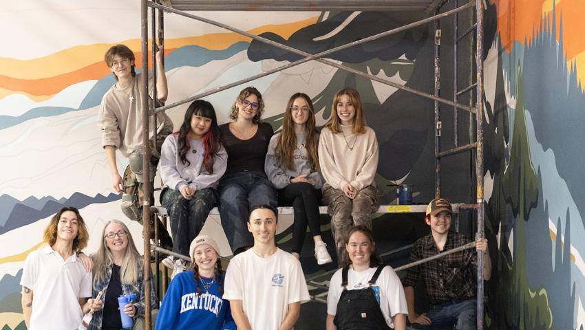 group of students pose in front of large mural in progress on building wall