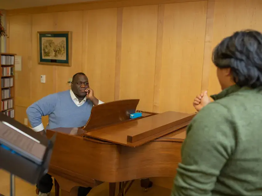 Image of Reggie Smith Jr. seated at a grand piano watching a student in the foreground during a vocal lesson.
