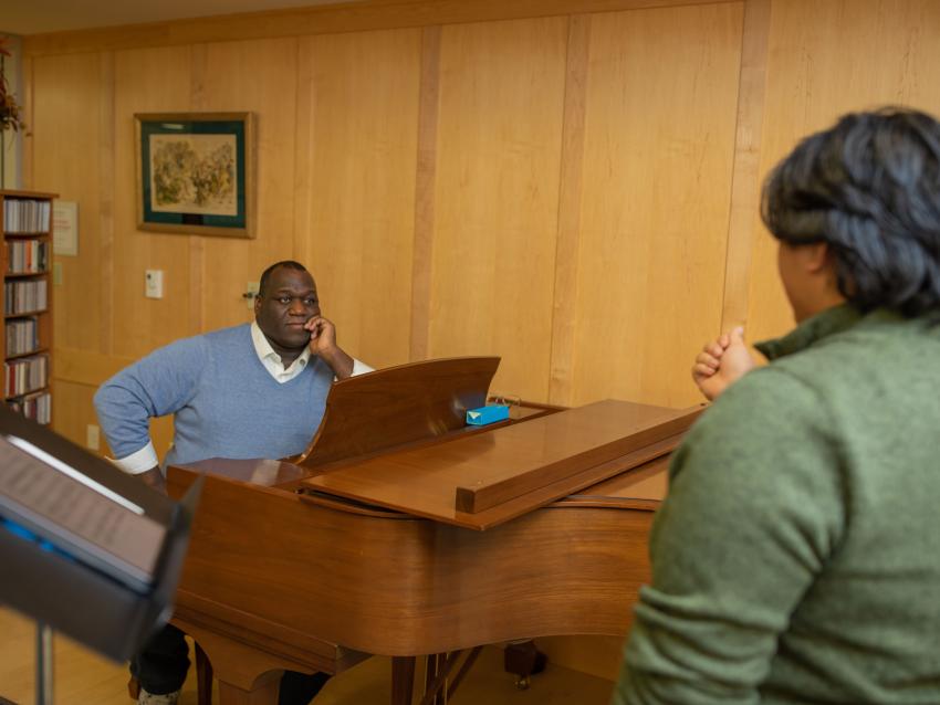 Image of Reggie Smith Jr. seated at a grand piano watching a student in the foreground during a vocal lesson.