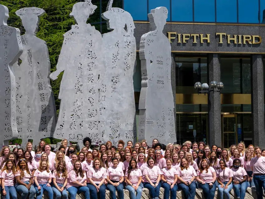 A large group of UK Treble Choir sits outside wearing pink tshirts in front of a large metal statue depicting silhouettes of female suffragettes, in front of the Fifth Third Bank building downtown Lexington  