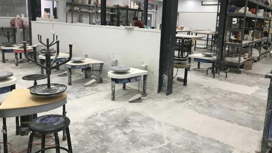 A wide view of the ceramics studio were rows of several throwing wheels are lined up together, shelves are along the back wall for works in progress, and more shelves farther away hold supplies.