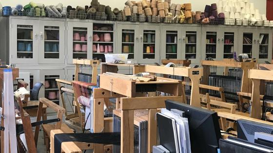Looking over a room packed with floor looms, many of which have works in progress warped on. Against the back wall are an extensive set of glass door cabinets packed with a rainbow of industrial sized cones of thread and yarn. More colors and sizes of cones are piled on top of cabinets.