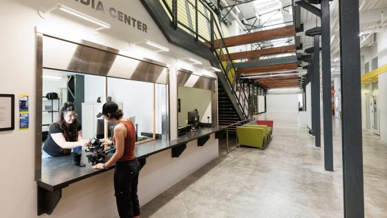 Two open counter bays sit under the main stairs of the Art and Visual studies building with "Media Center" printed above the first counter. A student worker at one counter assists another student in checking out some photography equipment.