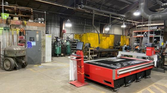 Interior of a large metal building. A bright red digital plasma cutting table is nearest the viewer; with many gas tanks, welders, tables, and other equipment to work with metal in the background.