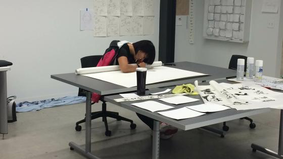 A studnet works on a large rolled out sheet of paper at a table in the middle of a white room. Taped up on the wall behind them are several sketches and iterations on designs.
