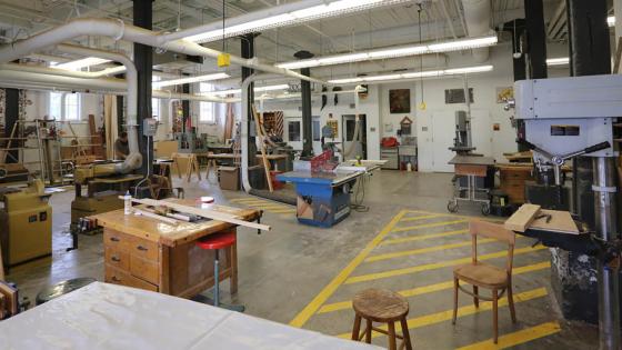 A huge warehouse-like room with copious ventilation ducts and lightning. A drill press is on the close right, and in the distance a tablesaw, planar, bansaw, jointer, and other heavy equipment can be seen. Yellow painted caution zones are marked generously around each piece of equipment.