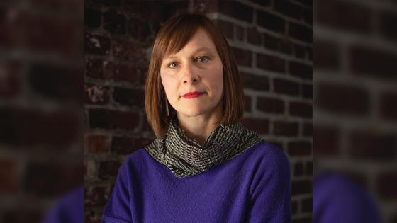 Image of Miriam Kienle, wearing a blue sweater with grey scarf against a brick wall