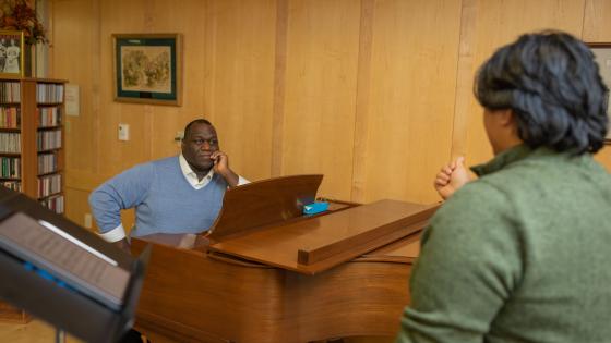 Image of Reggie Smith Jr. seated at a grand piano watching a student in the foreground during a vocal lesson.