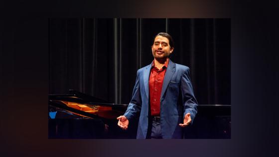 Image of Wagnor Pastor wearing a dark grey suit and red shirt performing onstage in front of a grand piano.
