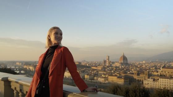 Film still of "Va Bene Così" a comedy short film written and directed by Peter Allen Stone that was filmed in Florence, Italy, woman in red coat stands on a bridge over looking Florence.