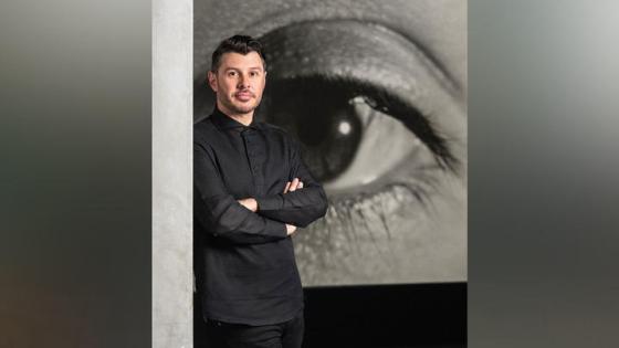 Image of Aaron Levi Garvey wearing a black shirt with his arms folded in front of a large sketch of an eye