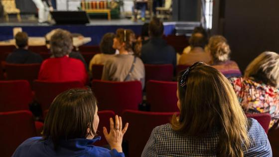 People sit in the audience of a small theatre facing away from camera.