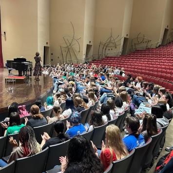 Photo taken from the recital hall seats facing the stage. A diverse and colorfully dressed body of student singers fill the first 4 rows of seats while director Beth Wilson speaks next to a concert piano on stage.