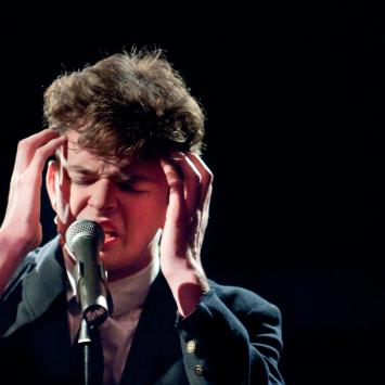 Student singing at a microphone wearing a blazer and white dress shirt, with hands on temples and eyes closed.