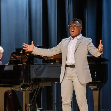CoryOn Brooks (2024) performing in front of a dark blue velvet curtain, with Professor Nan McSwain accompanying on a grand piano in the background. Photo by Sally Horowitz Photography.