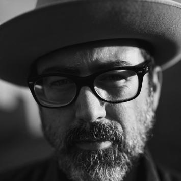 Black and white close up portrait of Brett Ratliff, bearded, with dark eye glasses and light fedora hat