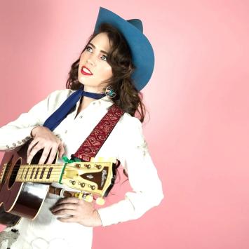 Image of Martha Spencer in white cowgirl outfit, blue cowboy had, holding a guitar, against a pink background