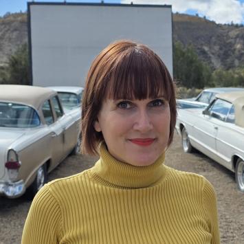 Lucy Jones, wearing a yellow ribbed turtleneck sweater, stands in the parking lot of a countryside drive-in movie theatre with retro and classic cars. Photograph by Mark Murray.