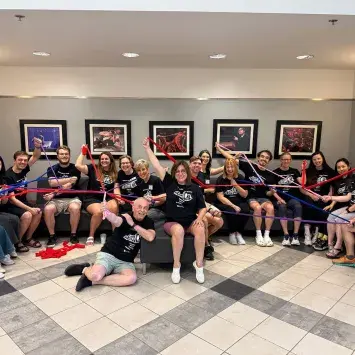 A class of 17 smiling students wearing matching t-shirts sit around a large couch area in the Guignol Theatre lobby. An assortment of performance photos hang on the wall behind them. Each of the students is holding one end of a stretchy colored ribbon in each hand while the ribbons criss-cross and twist around the group where other students to hold the opposite end.