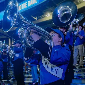 The Wildcat Marching Bands stands and plays in the bleachers of Kroger Field during a game with Notre Dame. Current uniforms have black pants, a UK blue and black jacket, with an embroidered KENTUCKY sash stitched across the front. Th band member in the foreground plays her silvery Euphonium, with other brass section members in the background playing a verity of instruments.