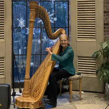 Elain Cook playing a large concert harp in front of a tall window at dusk decorated with snowflake ornaments.