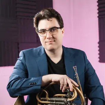 Scott Leger sits wearing a dark blue suit in a light pink room with black panels of acoustic foam and holds his french horn in his lap.