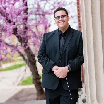 Portrait of Alex Mondragon in a black suit leaning against a carved column with a brilliant purple blooming tree in the background. He casually holds a conductor's baton in his clasped hands.