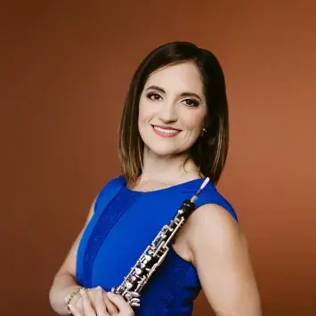 Headshot of ToniMarie Marchioni wearing a blue dress in front of a burnt orange background while holding her oboe.