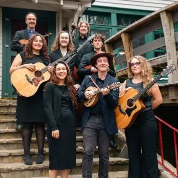 Eight members of the band stand together smiling with various instruments on rustic stone steps in front of a historic building.