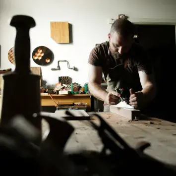 Daniel Graham in his workshop, hand planing a piece of wood. An assortment of tools, pieces in progress, and finished works can be seen around the room.