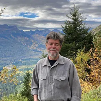 Jeff Warner stands on a mountain with a vibrant valley an other mountains spread out in the distance behind him.