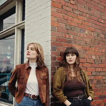Two women look casually into the distance leaning against the corner of a small town brick andf glass storefront.