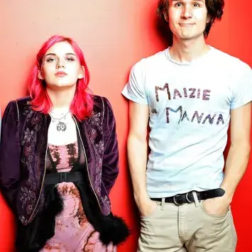 Two eclectic young starts stand in front of a vibrant grapefruit colored wall. Maizie Manna, with neon pink hair and dark jacket, stands on the left. A band mate with a shirt that reads "Maizie Manna" stands on the right.