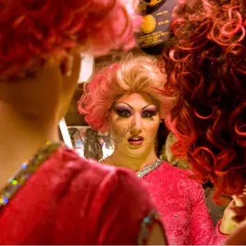 A performer in a bright pink costume and curly pink wig applies makeup while looking into a mirror backstage. The image is framed through the reflection, showing detailed stage makeup and costume embellishments. Another red-and-black curly wig appears in the foreground, partially surrounding the mirror. The warm lighting and dressing-room setting highlight preparations for a theatrical performance.