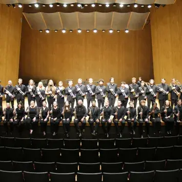 39 students plus Professor Jason Dovel are in black formal wear on the softly lit Singletary Recital Hall stage. Half of them are standing in a row behind the other half who are sitting on the edge of the stage.