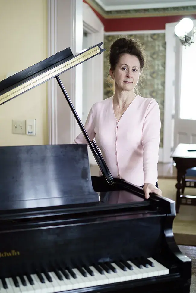 Irina Voro standing by a piano in a softly lit room. Photo courtesy of Changpeng Ti.