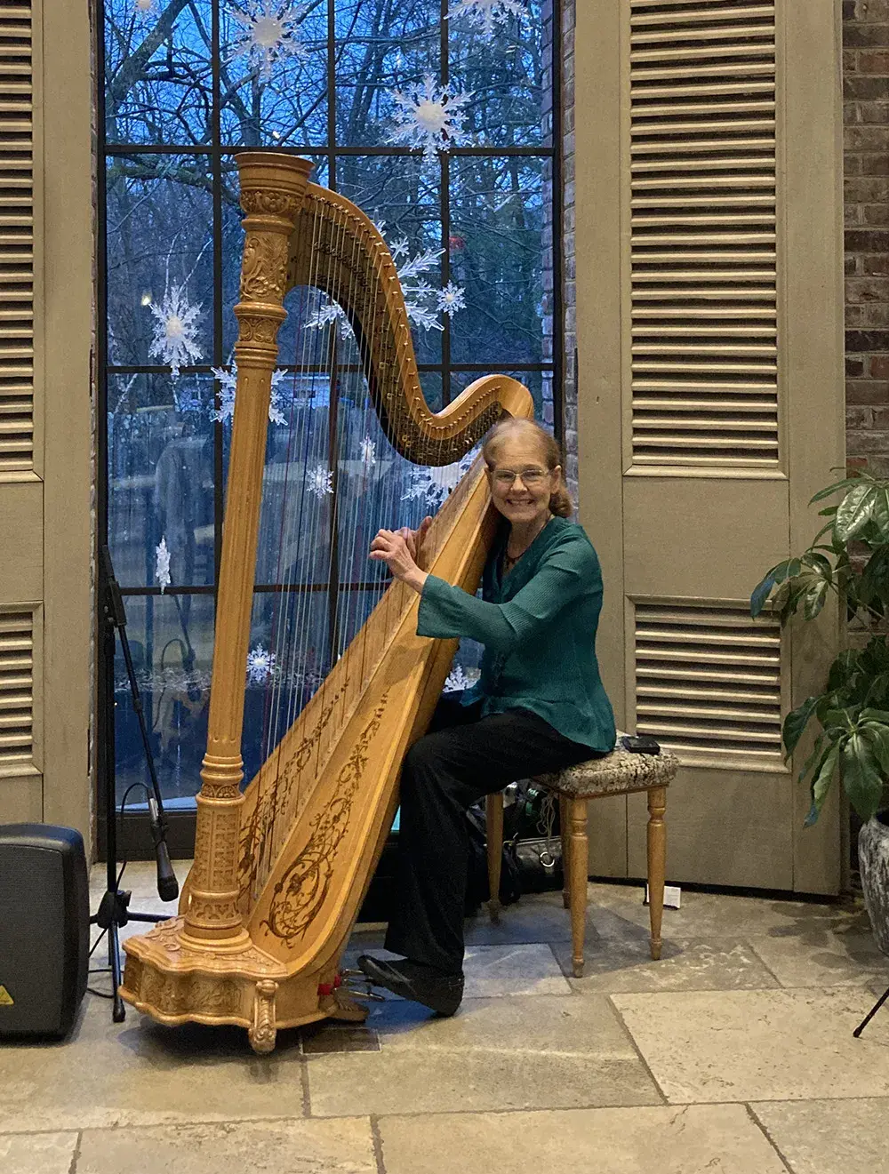 Elain Cook playing a large concert harp in front of a tall window at dusk decorated with snowflake ornaments.