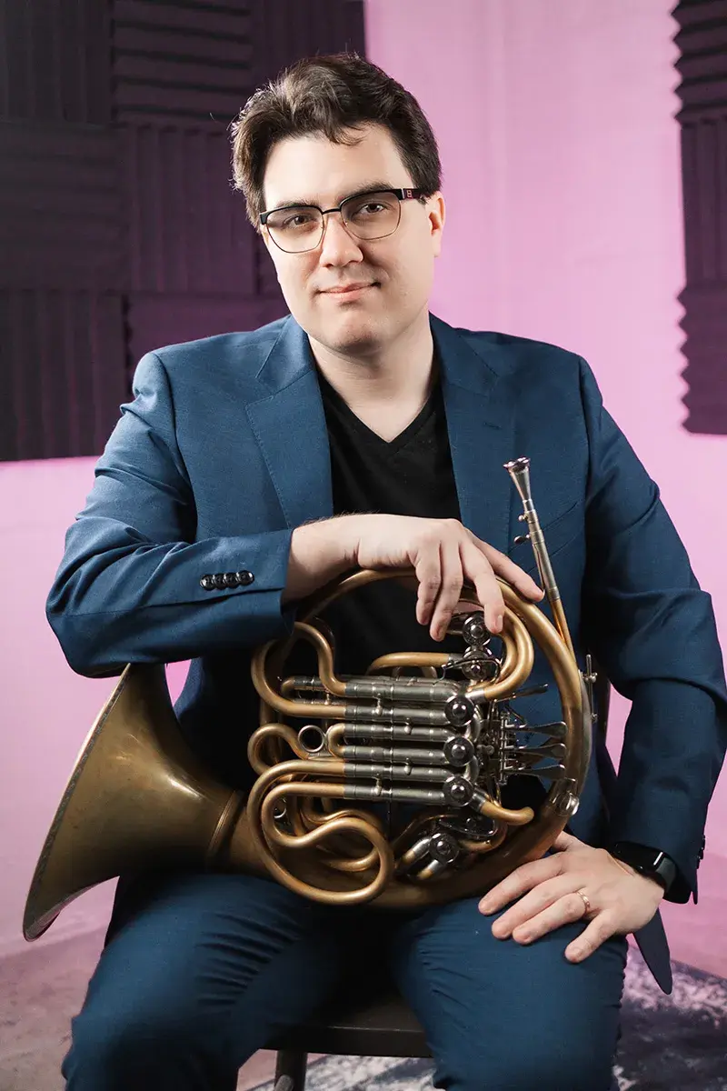 Scott Leger sits wearing a dark blue suit in a light pink room with black panels of acoustic foam and holds his french horn in his lap.