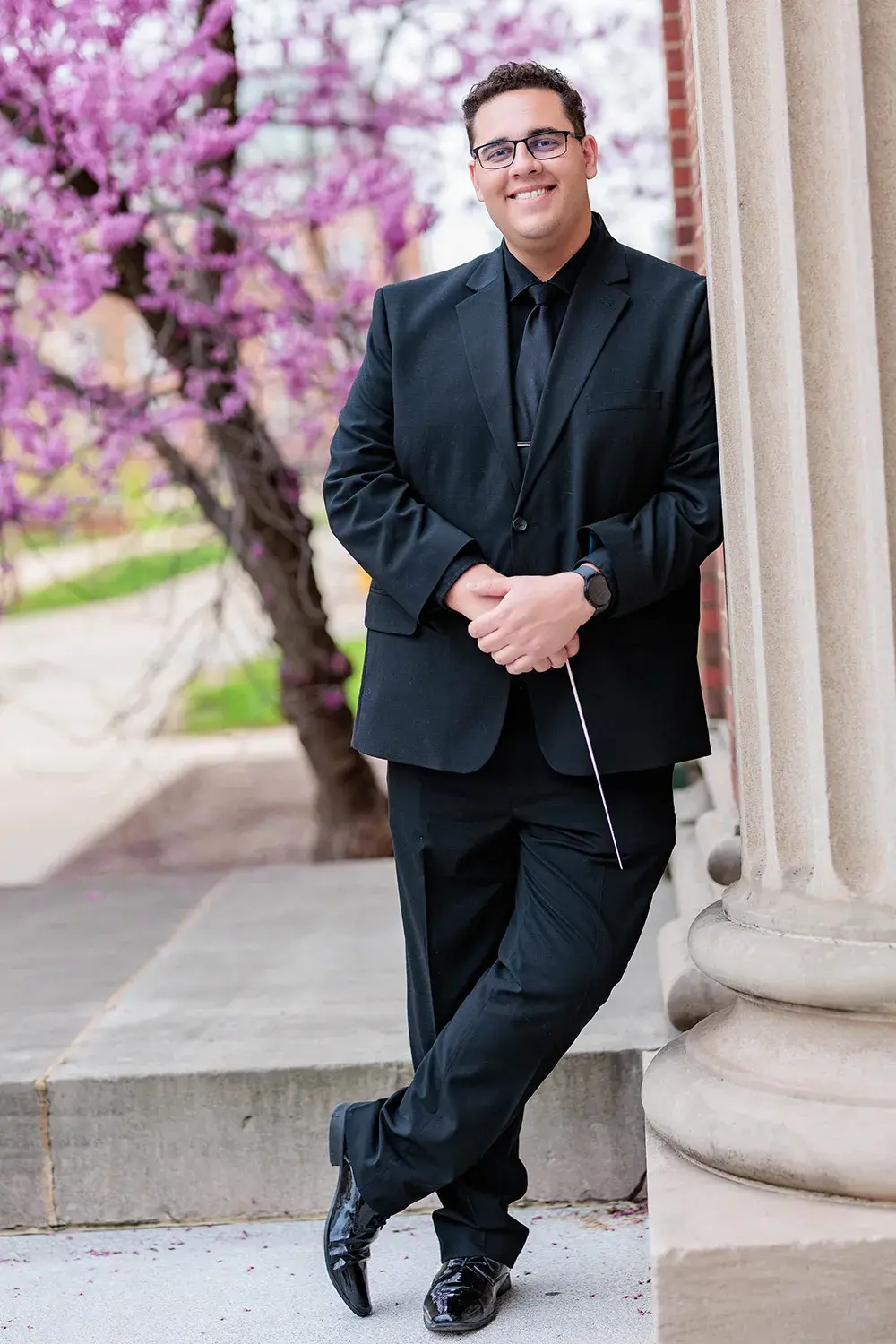 Portrait of Alex Mondragon in a black suit leaning against a carved column with a brilliant purple blooming tree in the background. He casually holds a conductor's baton in his clasped hands.