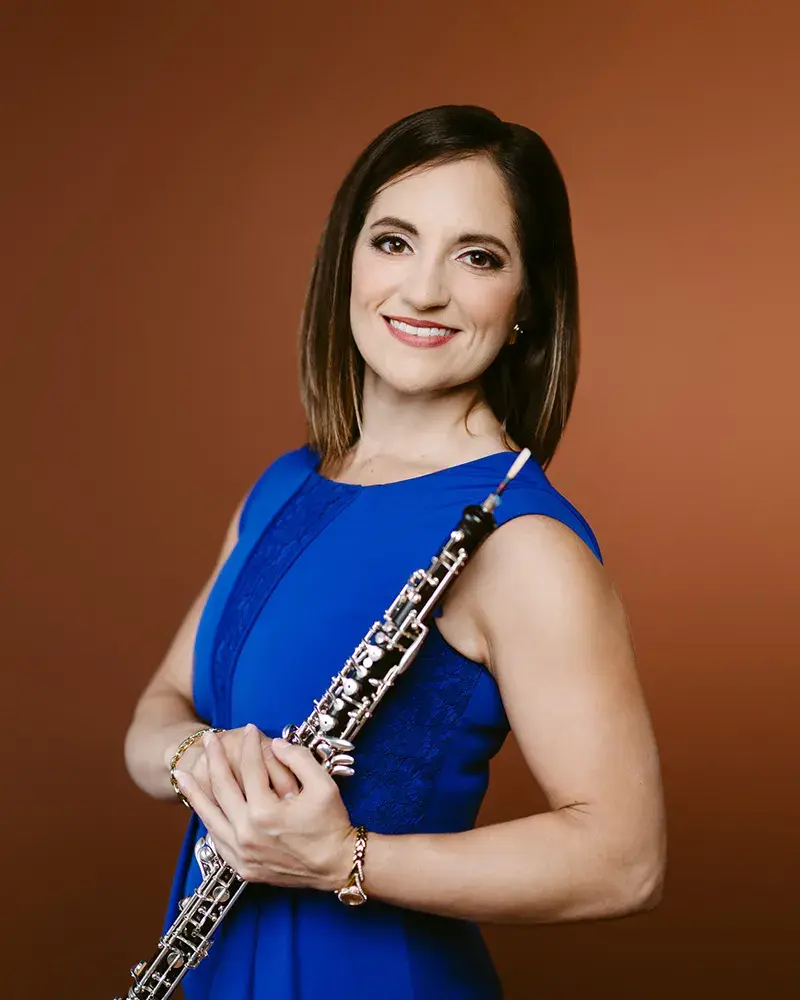 Headshot of ToniMarie Marchioni wearing a blue dress in front of a burnt orange background while holding her oboe.